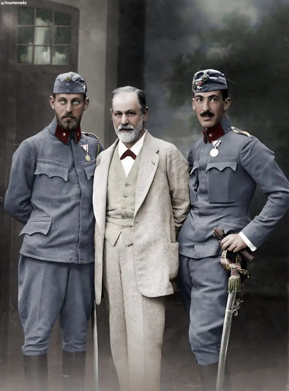 Three men pose in a vintage photograph: a central older man in a light-colored suit and tie flanked by two younger men in…