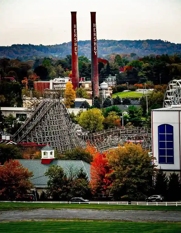 A scenic view of the Hershey Park amusement park featuring two tall red smokestacks with 'HERSHEY' written vertically in…