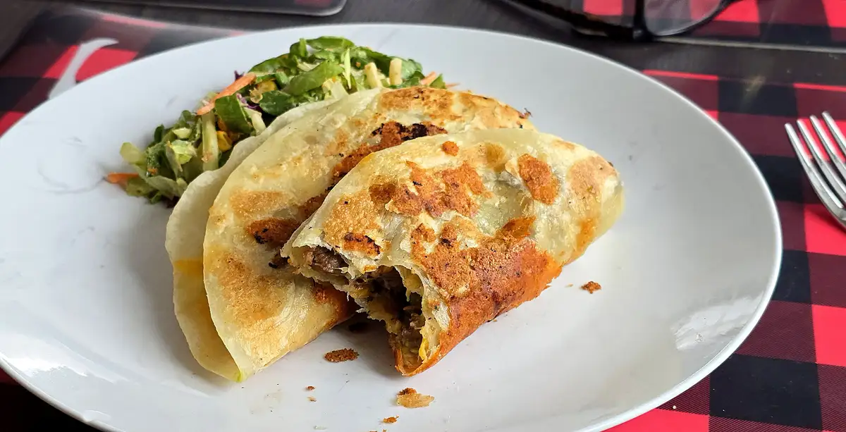 A white plate holds two golden-brown, crispy-edged fried dumplings, one broken open to reveal meat filling, alongside a…