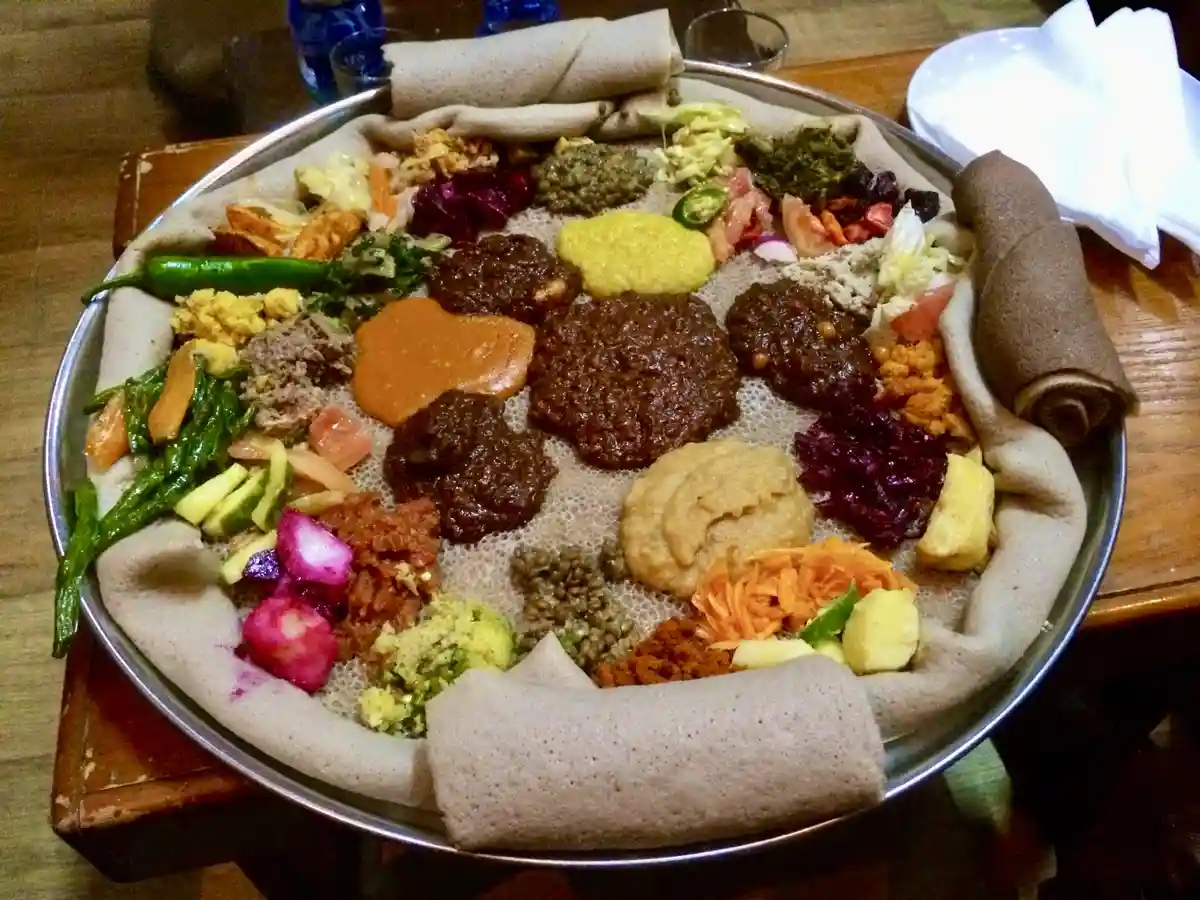 A large metal platter holds a traditional Ethiopian meal, featuring a spongy, sourdough flatbread called injera spread…