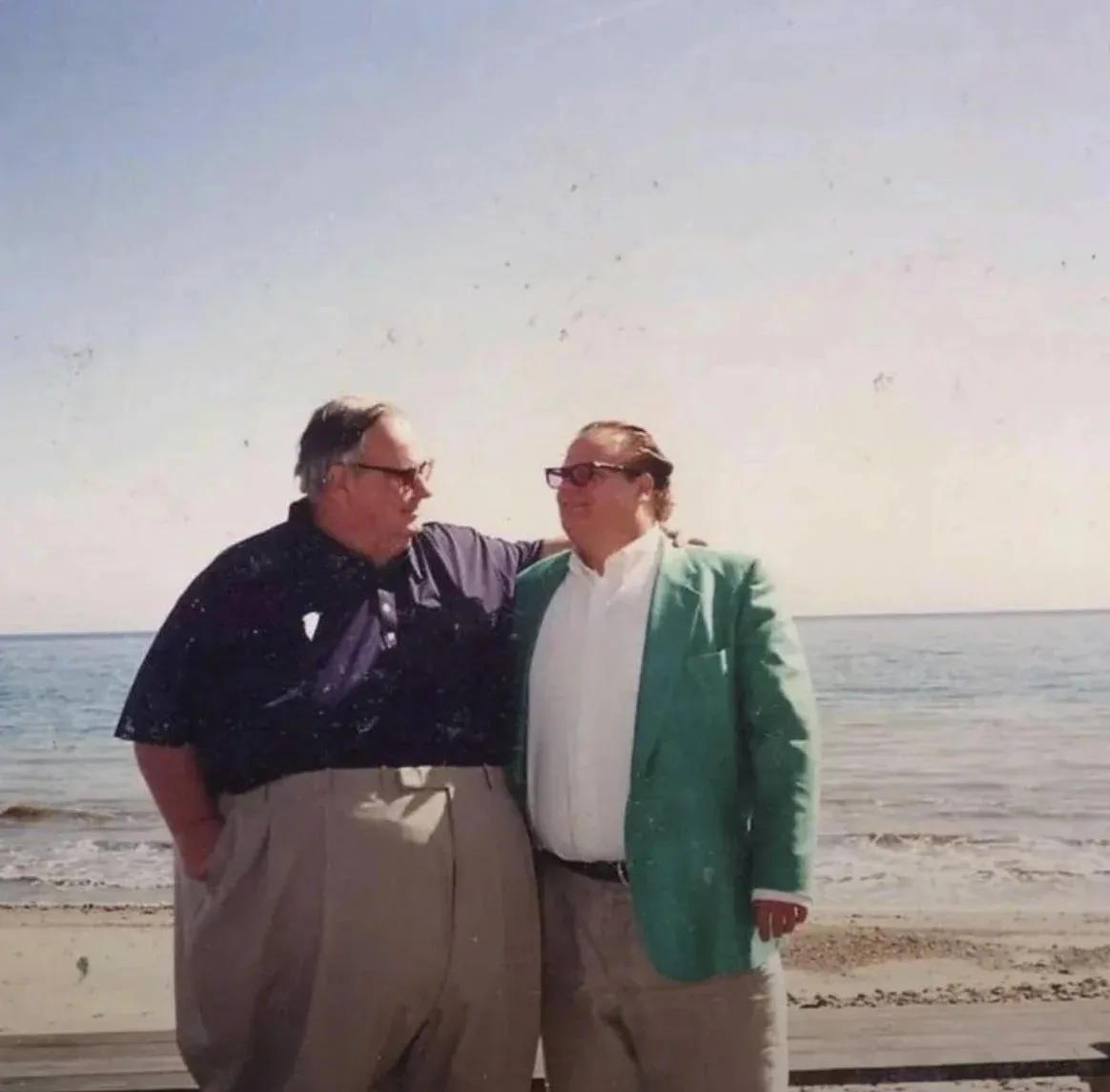 Two men stand on a beach with the ocean behind them; one wears a dark shirt and khakis, the other wears a bright green…