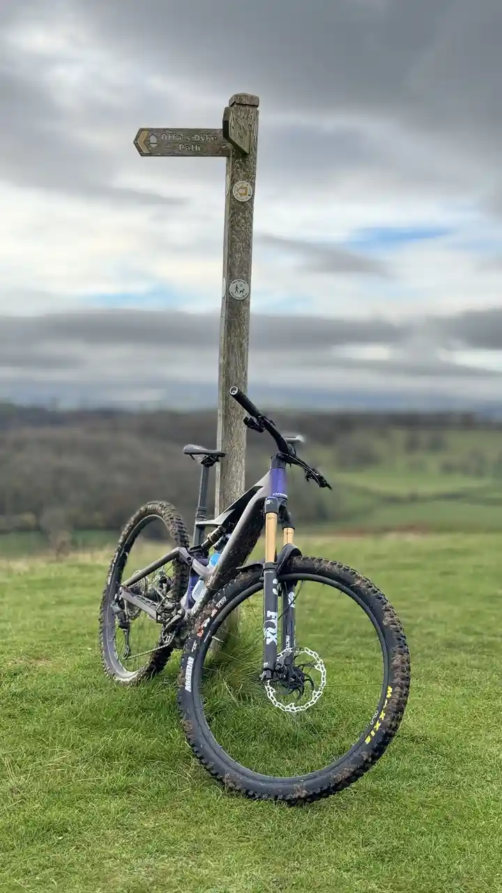 A muddy mountain bike is leaning against a wooden trail signpost labeled 'Offa's Dyke Path' in a grassy field under a cloudy…