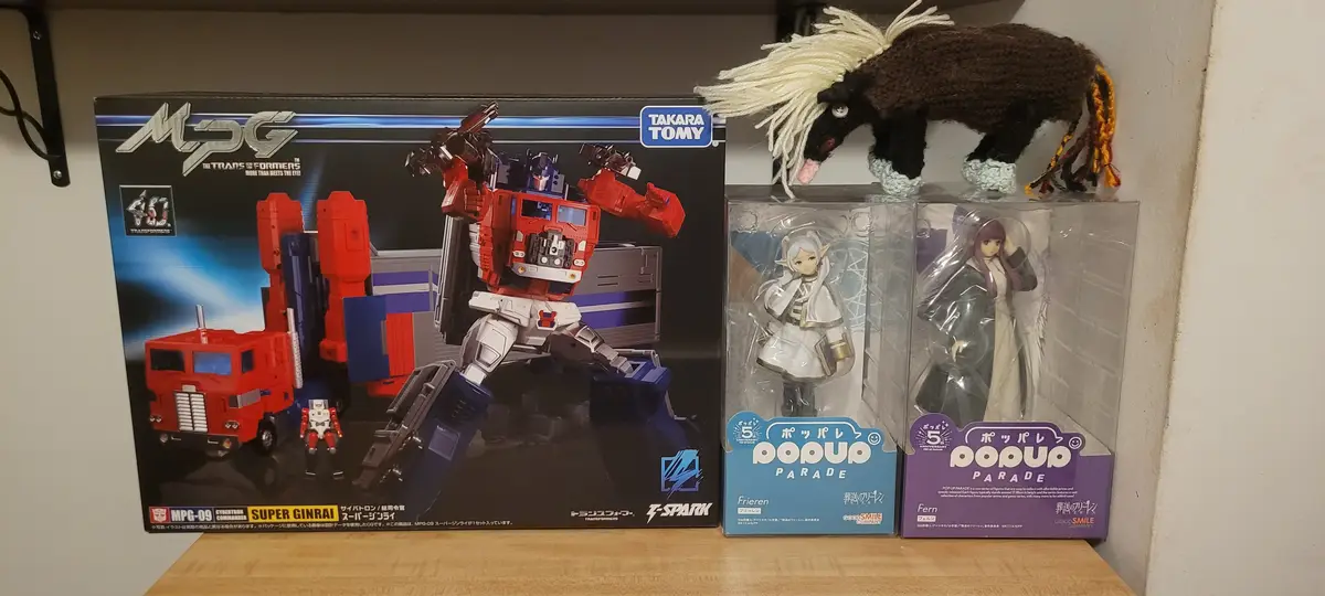 A shelf displays a large Transformers toy box for 'Super Ginrai' next to two Pop Up Parade anime figures of Frieren and…