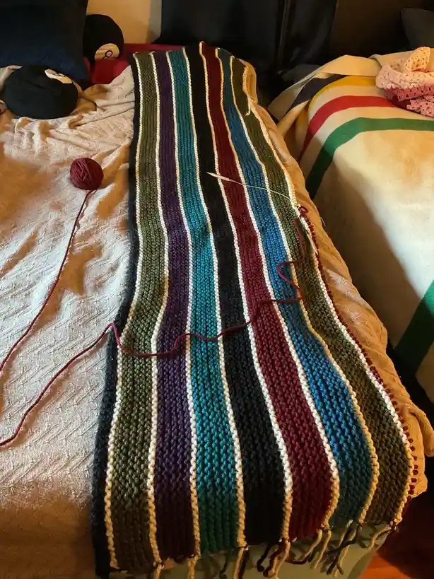 A colorful, striped knitted blanket with a red yarn ball and knitting needle rests on a bed, with the overlaid text reading…