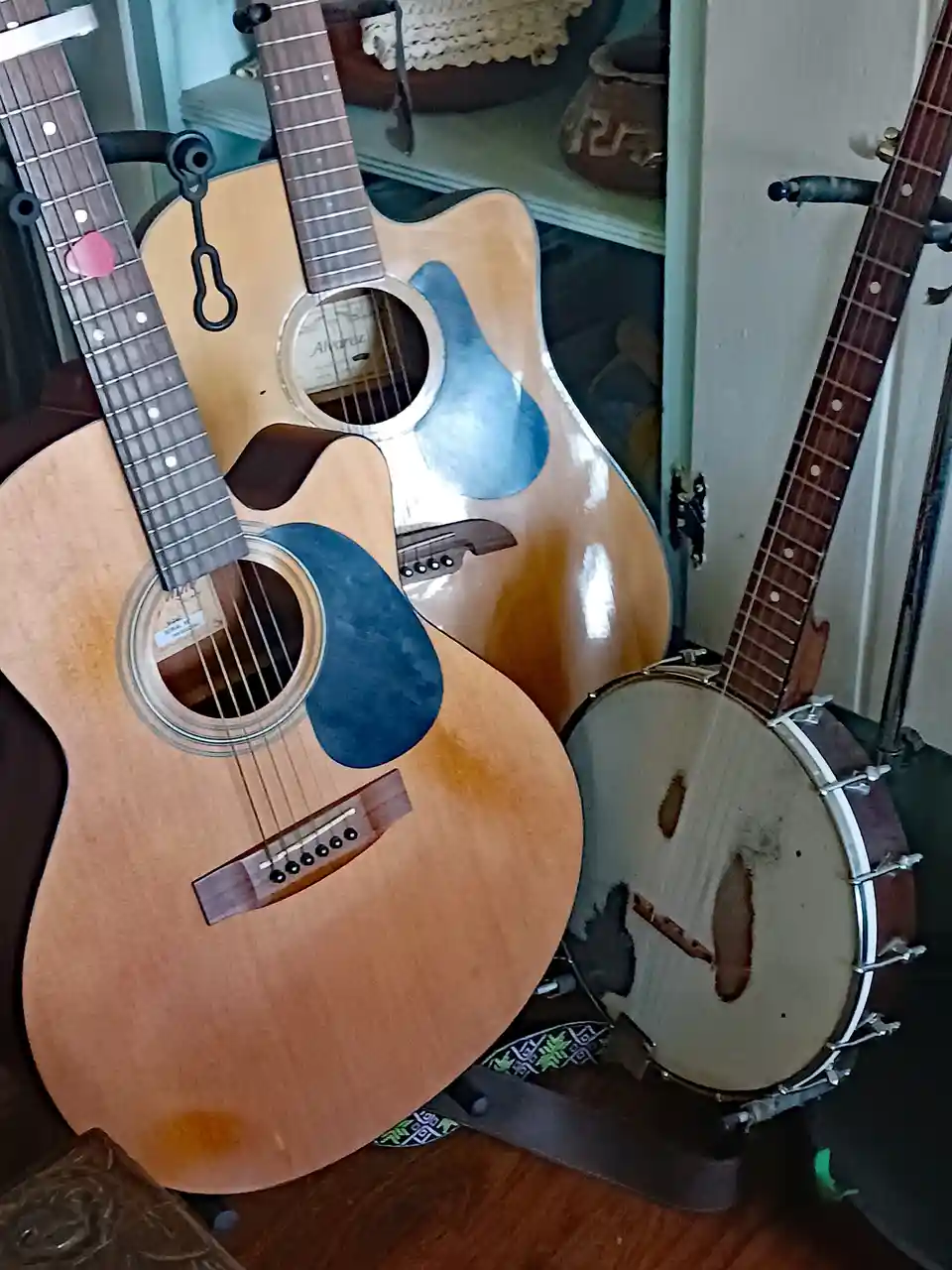 Two acoustic guitars with blue pickguards and a banjo with a worn head are leaning against each other on a wooden floor…
