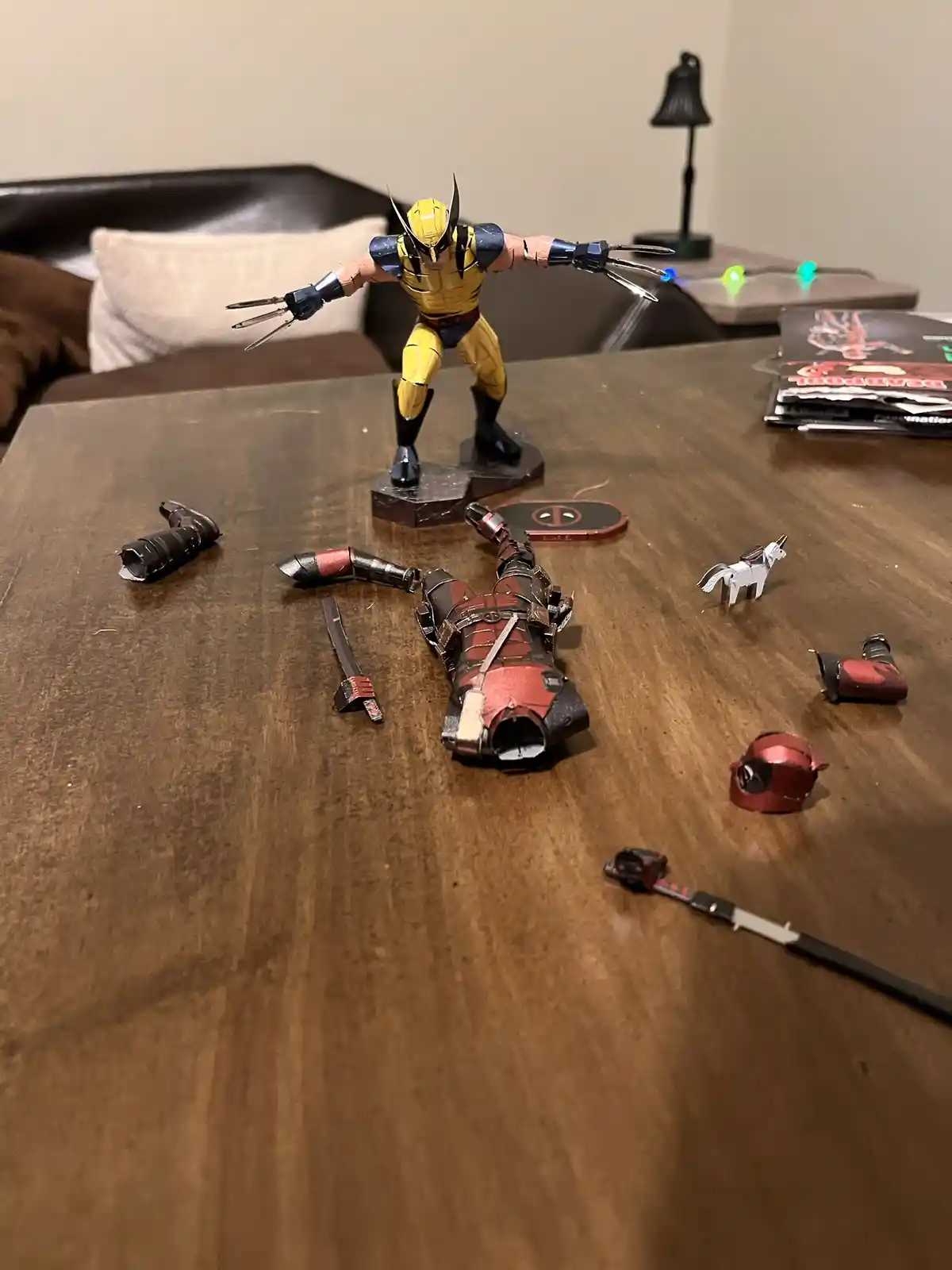 A Wolverine action figure stands victorious on a wooden table, facing a disassembled Deadpool figure whose parts are…