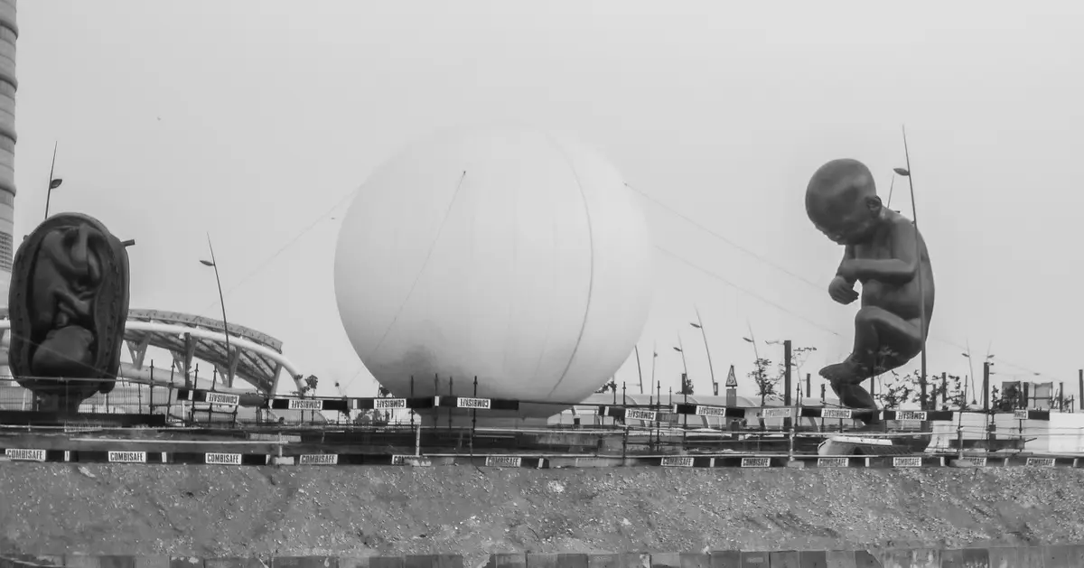 A black-and-white photo shows three large sculptures at a construction site: a fetal figure in an amniotic sac on the left…