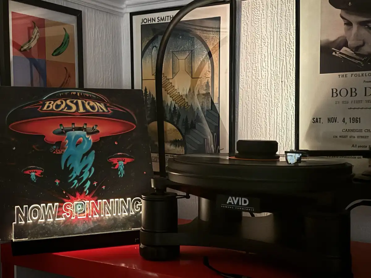 A vinyl record player sits on a red surface with the album cover for Boston's 'Now Spinning' album prominently displayed in…