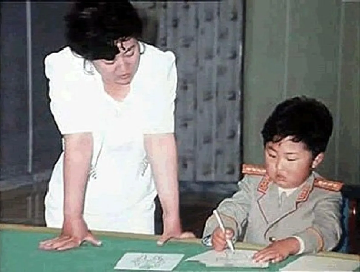 A woman in a white shirt leans over a green table, watching a young boy in a military-style uniform write with a pen. The…