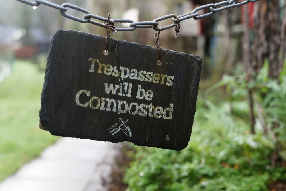 A dark, rough-hewn stone sign hangs from a metal chain in a garden setting, with the text 'Trespassers will be Composted'…