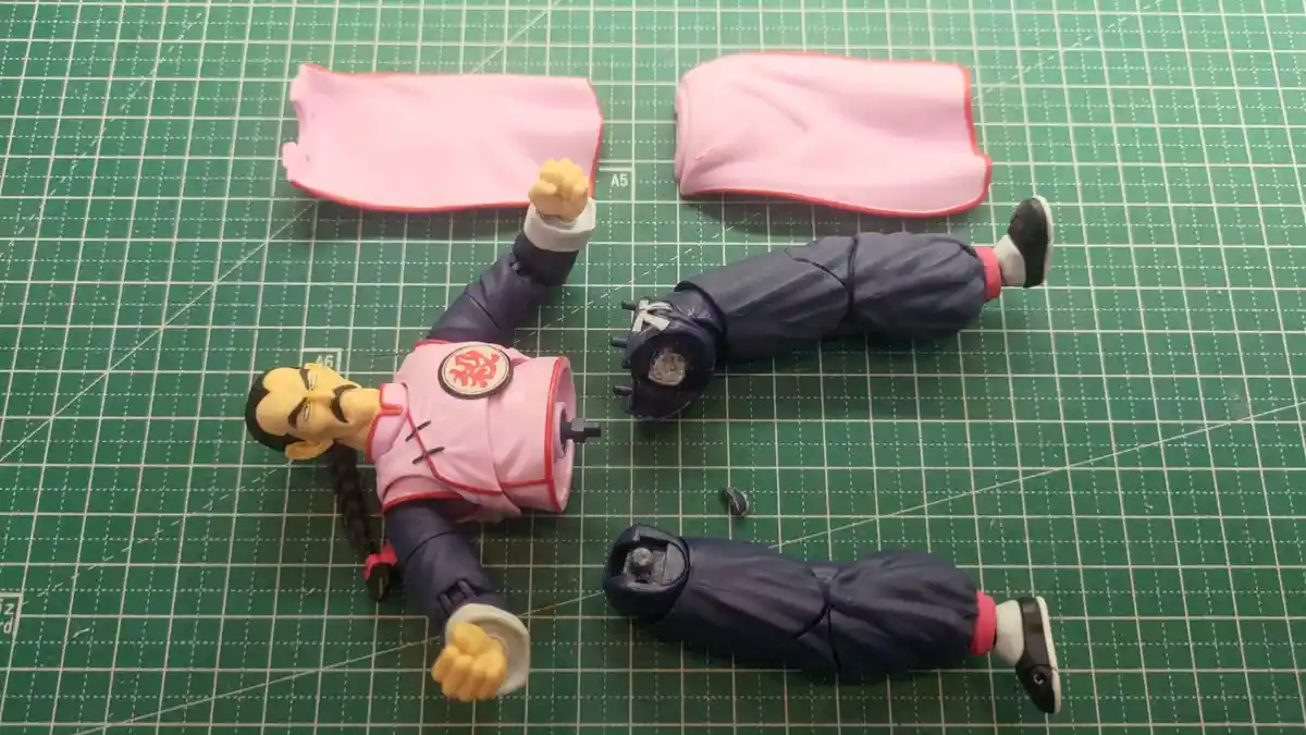 A broken action figure of a character with a pink top and dark pants lies on a green cutting mat, with its arms, legs, and…