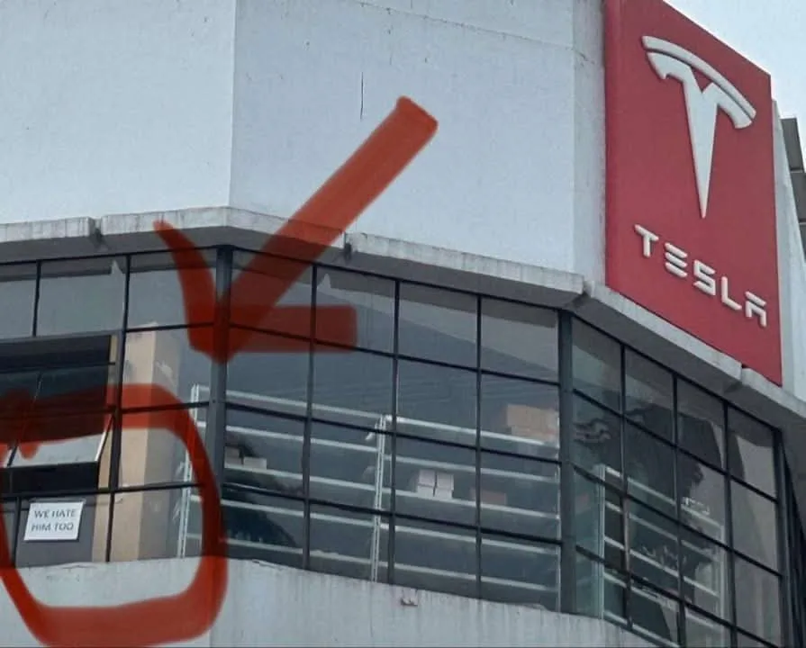 A photo of a Tesla building with a large red spray-painted arrow pointing to a small white sign on the wall that reads 'WE…
