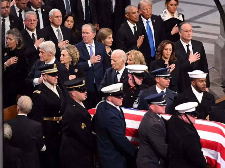 A solemn photo of U.S. political leaders and military personnel standing in a line during a funeral, with many placing their…