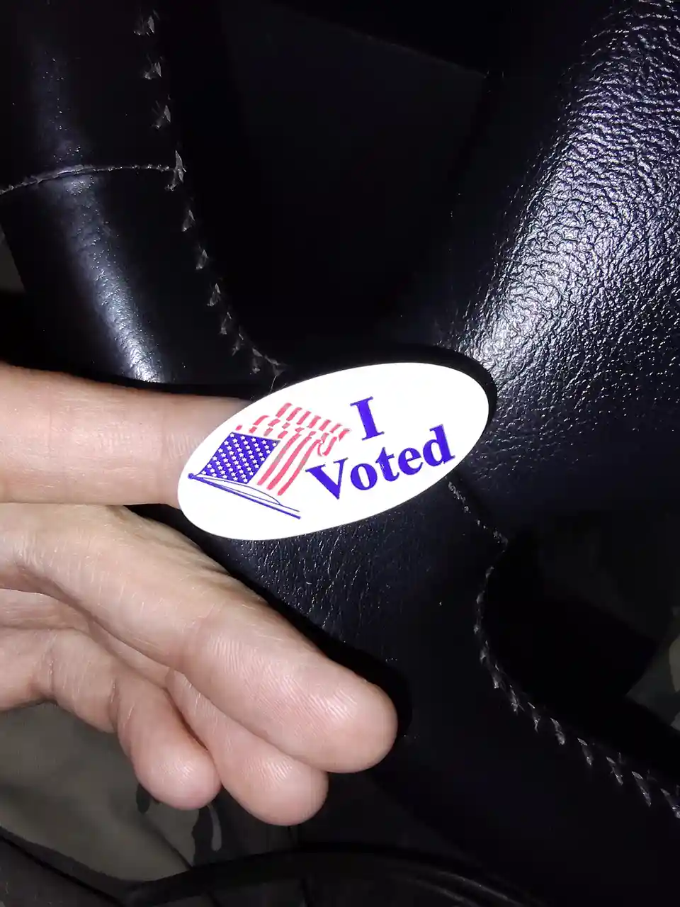 A close-up photo shows a person's finger holding a white oval sticker with blue text reading 'I Voted' and a small American…