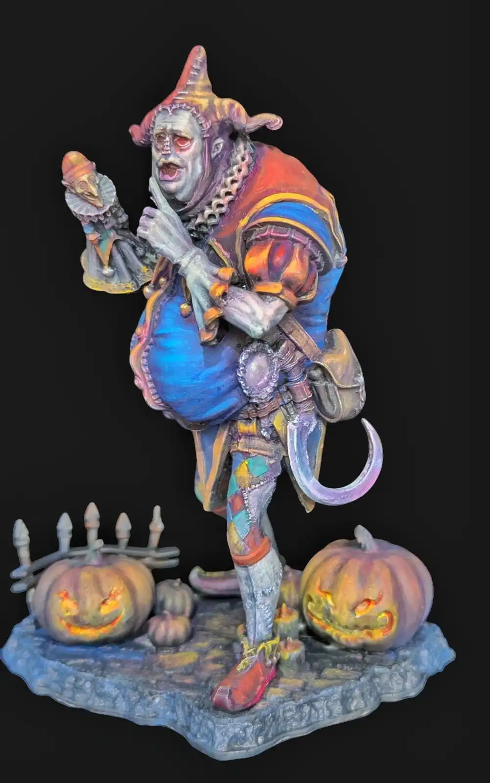 A painted figurine of a grotesque, skeletal jester with a pointed hat, holding a small puppet, standing on a base with two…