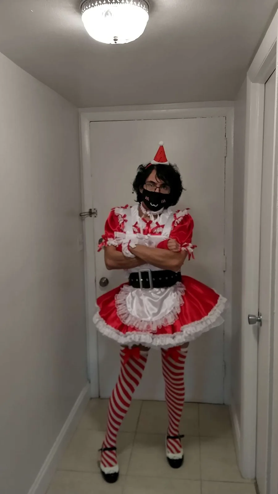 A person stands in a narrow hallway wearing a red and white Santa-themed maid outfit with striped stockings, a black mask…
