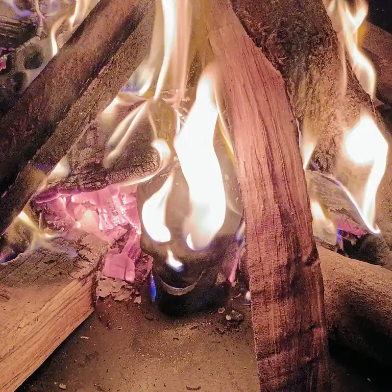 A close-up photo of a wood fire with flames licking up the sides of logs and glowing embers below. A small, dark…