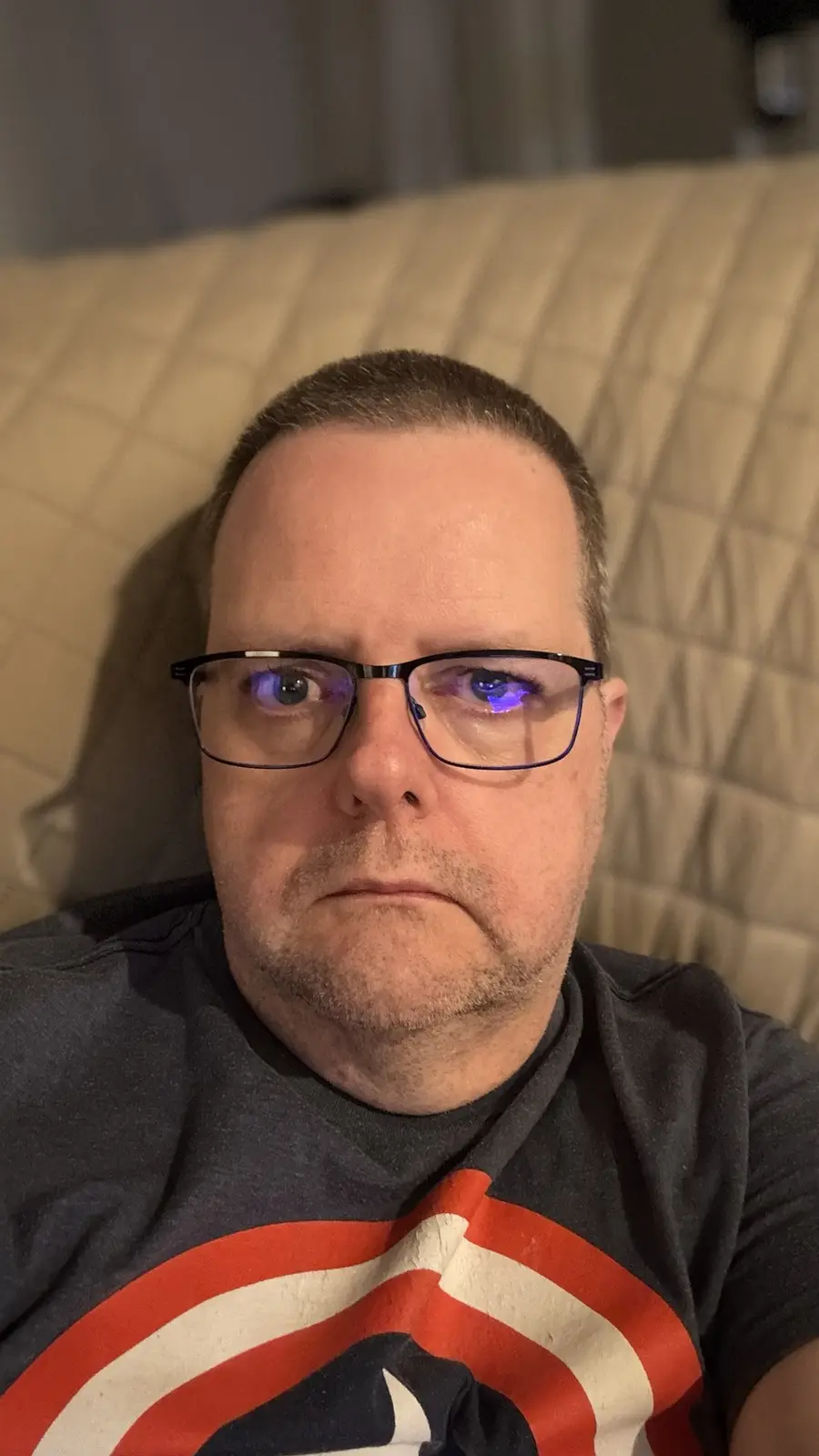 A man with glasses and a Captain America t-shirt sits on a couch with a tired, annoyed expression. His eyes reflect blue…