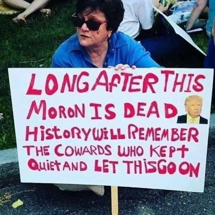 A person wearing sunglasses and a blue shirt holds a handmade protest sign with red handwritten text. The sign reads 'LONG…