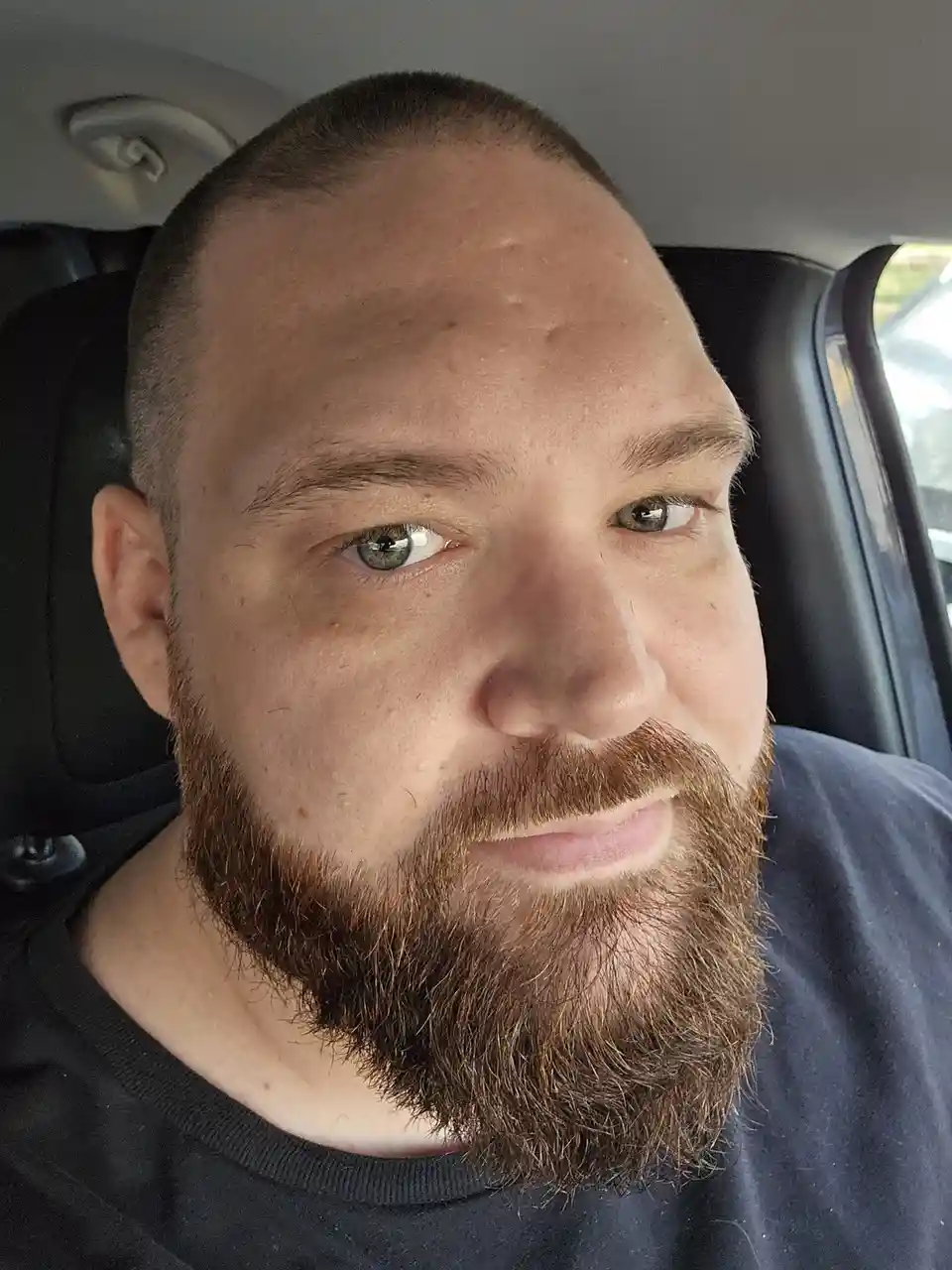 A close-up selfie of a man with a short buzz cut and a full brown beard, sitting inside a car. He is looking directly at the…