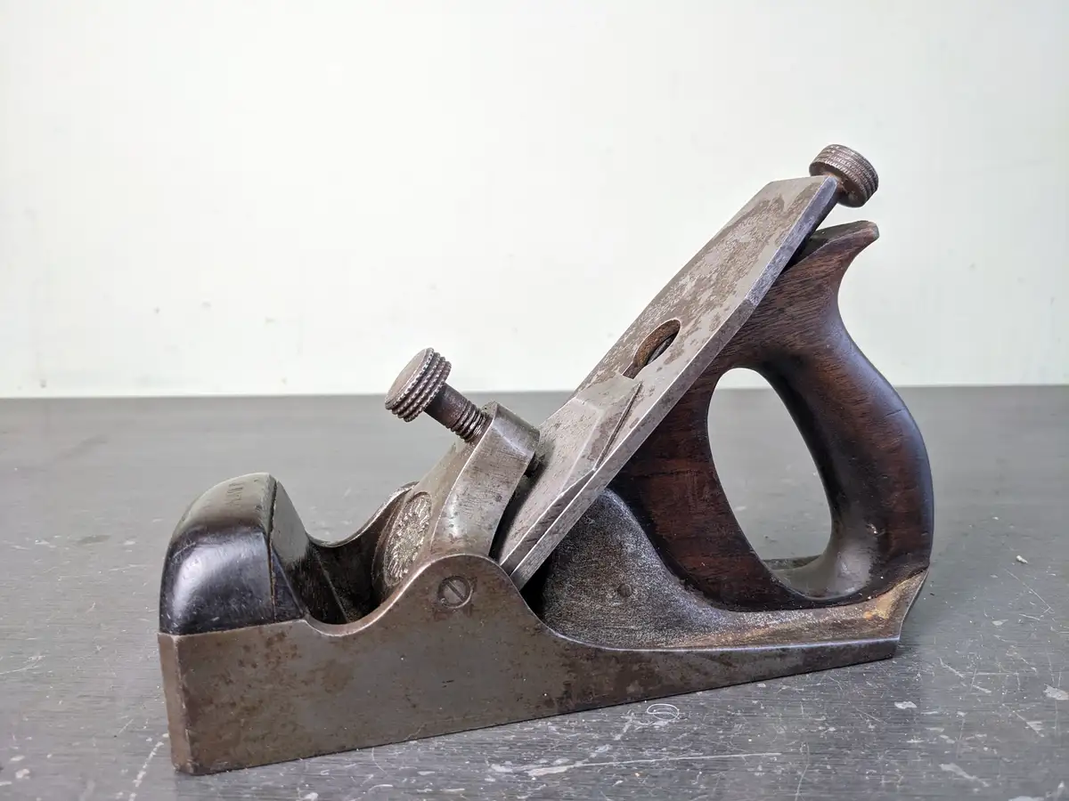 A well-worn, vintage hand plane with a dark wooden handle and metal body rests on a scratched metal workbench against a…
