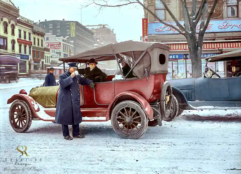 A vintage colorized photo shows a man in a police uniform drinking from a mug while standing beside a red early 20th-century…
