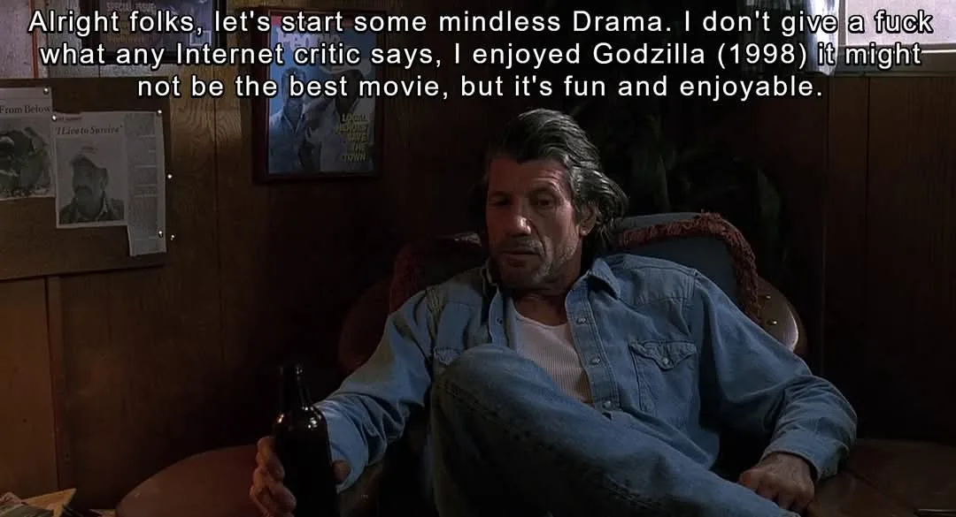 A man with graying hair and a beard, wearing a denim shirt and jeans, sits in a worn armchair holding a beer bottle. Behind…