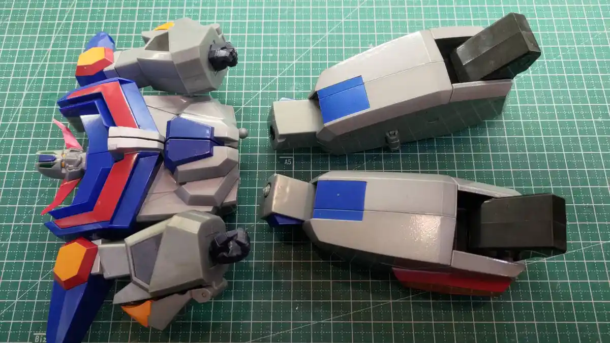 A partially assembled robot toy with a colorful blue, red, and gray body sits on a green grid mat, while its two detached…