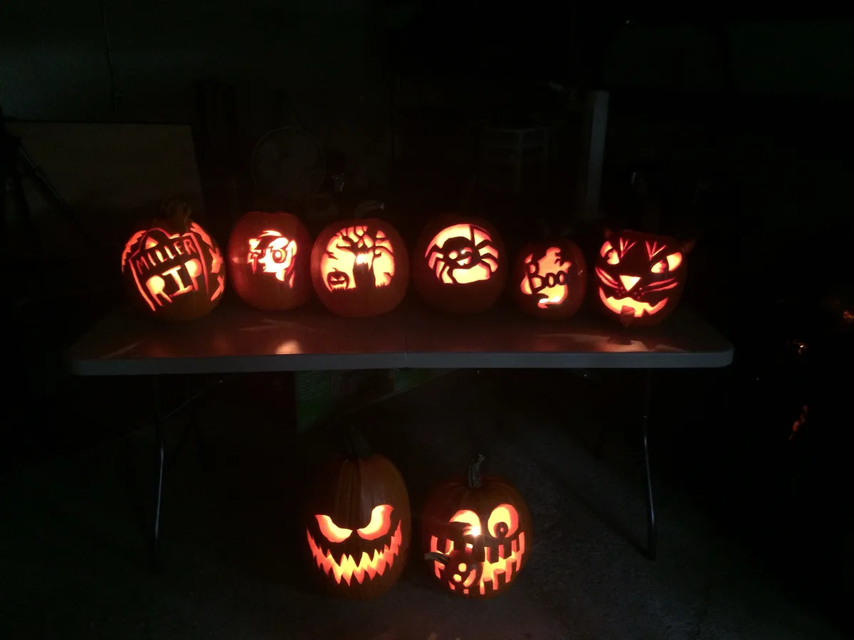 A row of seven intricately carved, glowing jack-o'-lanterns sits on a table in a dark room, each featuring unique designs…