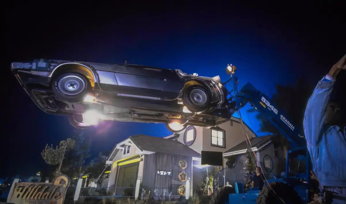 A black classic car is suspended upside down by a crane at night, with a person in a denim jacket raising their arm in the…