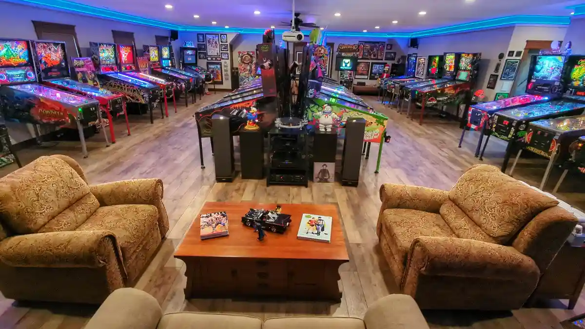 A wide-angle photo of a lavish, brightly lit basement filled with dozens of colorful, illuminated pinball machines arranged…