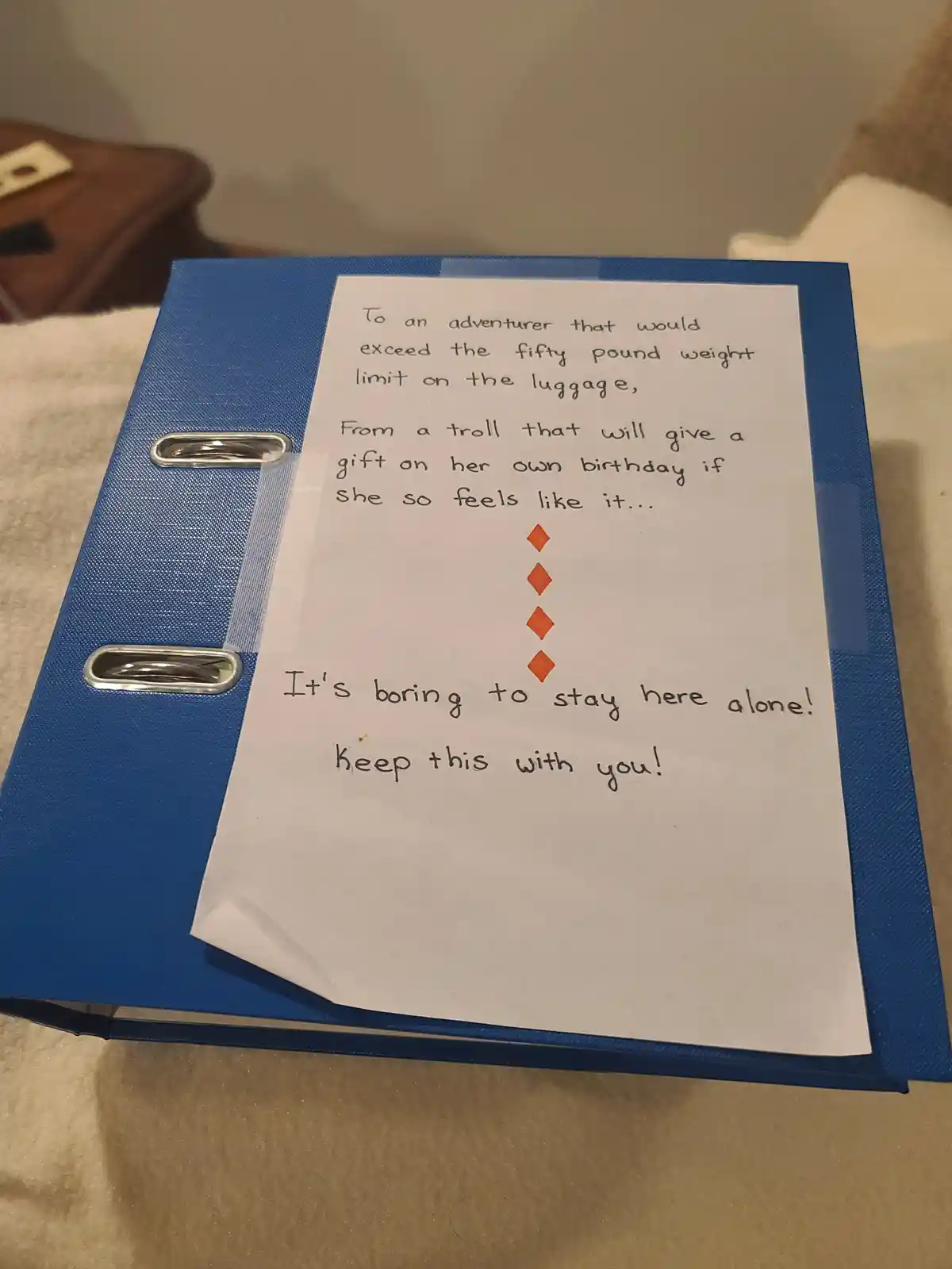 A handwritten note taped to a blue binder reads a playful, sarcastic message addressed to an adventurer who exceeds luggage…