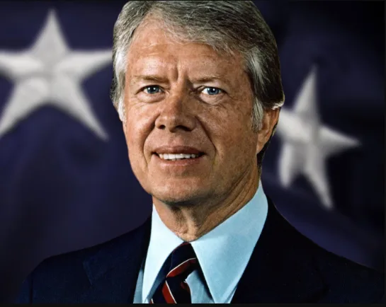 A formal portrait of former U.S. President Jimmy Carter, smiling, wearing a dark suit, light blue shirt, and red-and-black…