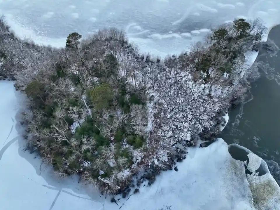 An aerial view of a small, snow-covered wooded island surrounded by frozen water with visible cracks and ice patterns. The…