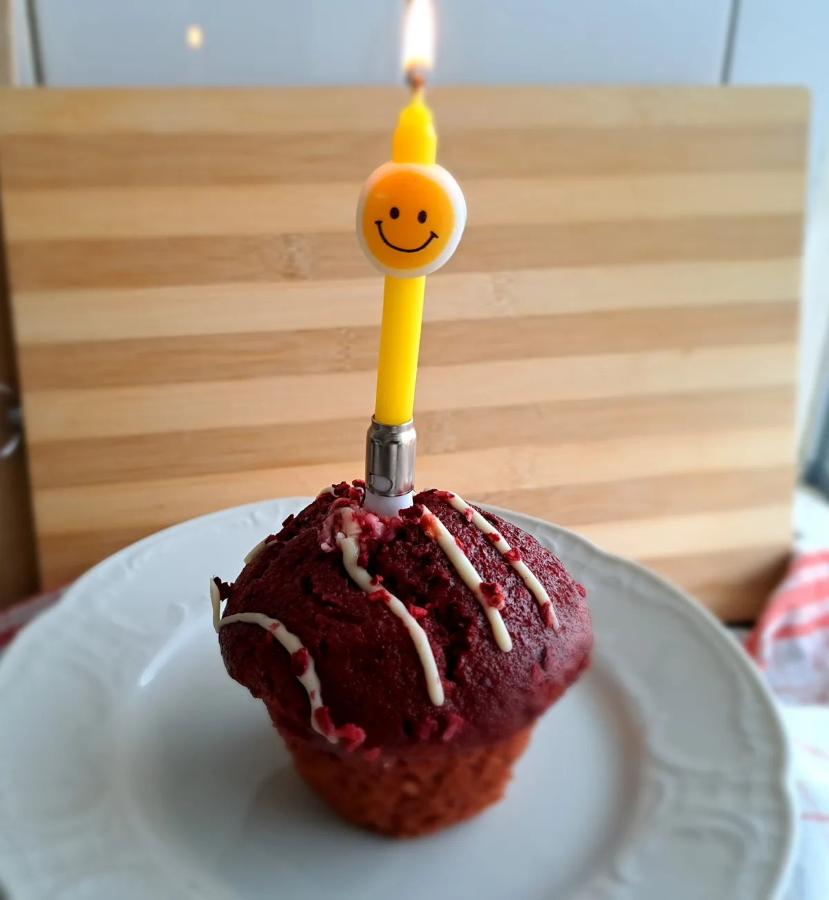 A red velvet cupcake with white icing drizzles sits on a white plate, topped with a lit yellow birthday candle featuring a…