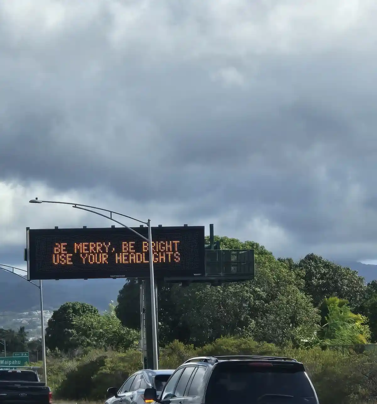 An electronic highway sign displays the message 'be merry, be bright use your headlights' in orange text against a dark…
