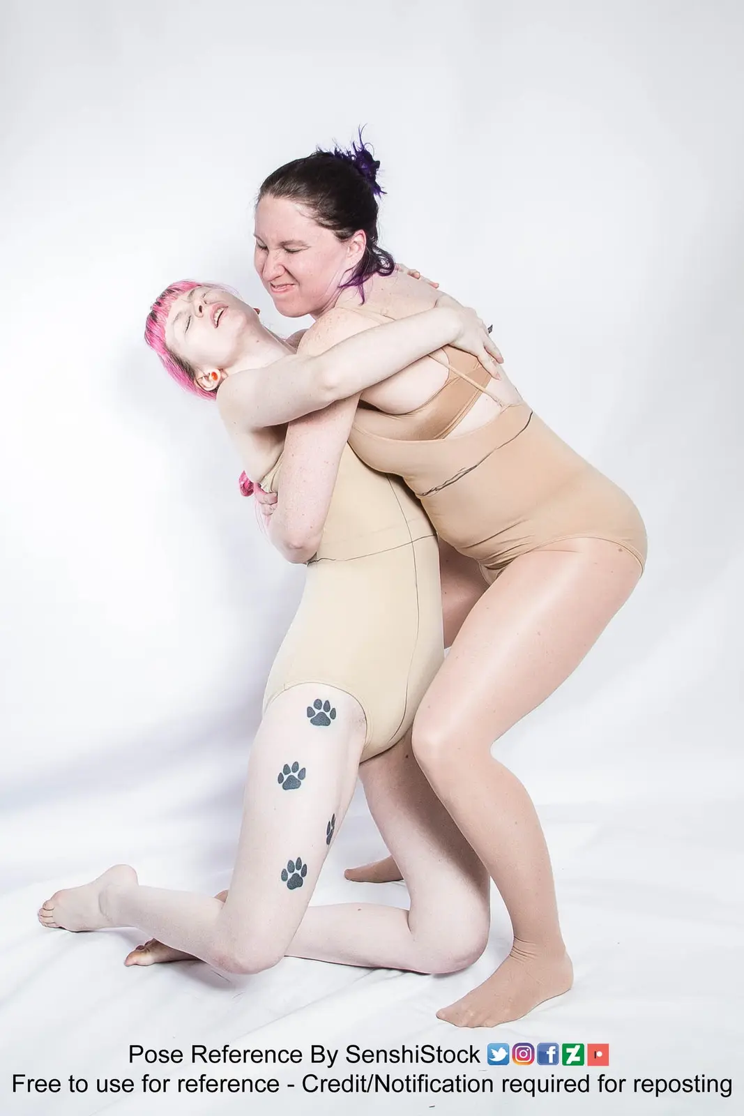 Two women in beige bodysuits pose against a white background; one kneels while the other straddles her, both smiling. The…