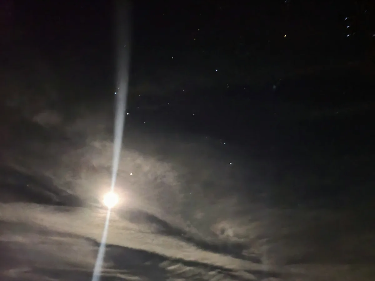 A night sky with stars and clouds, featuring a bright light source with a prominent lens flare extending vertically. The…