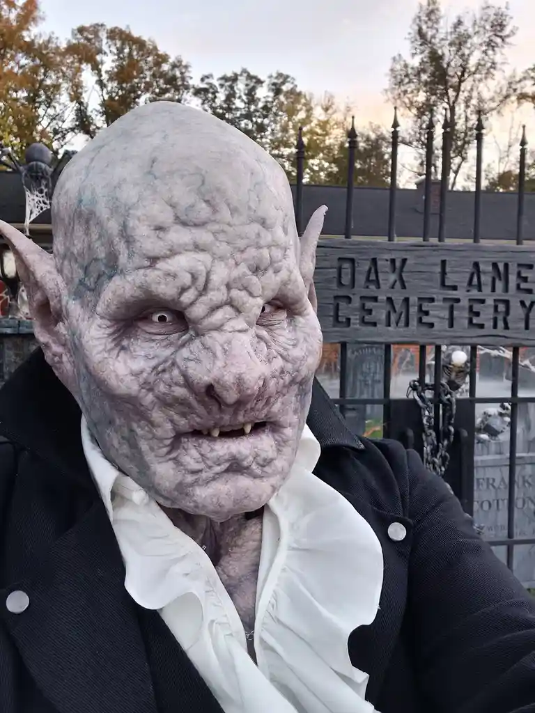 A person in a detailed, wrinkled, grey-skinned vampire costume with pointed ears and fangs stands in front of a black metal…