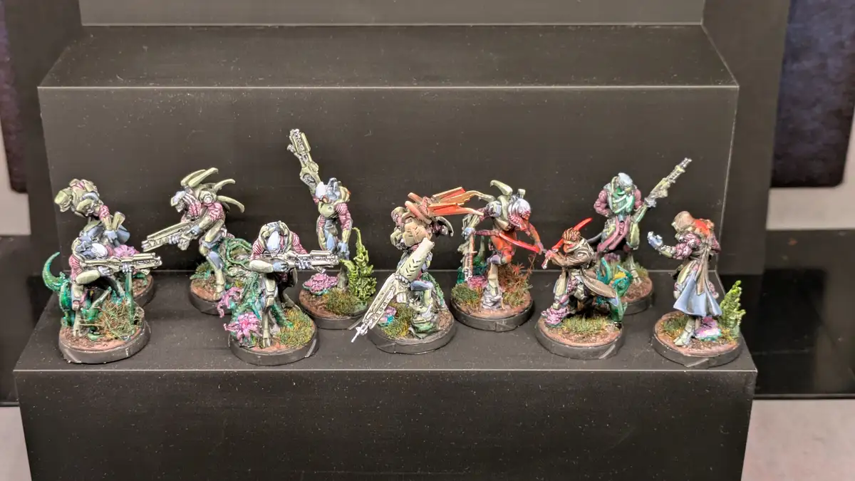 A row of eight painted miniature figurines, likely from a tabletop wargame, are displayed on a black shelf. Each figure is…