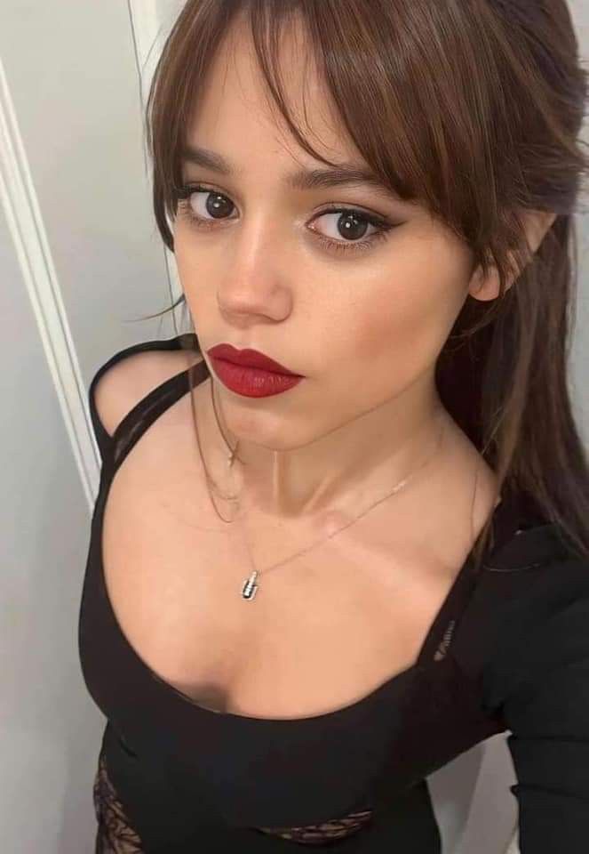 A woman with brown hair and red lipstick looks directly at the camera with a skeptical expression while wearing a black top…