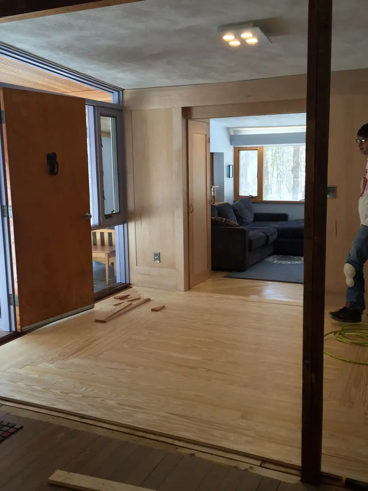 A person stands in a partially renovated room with light wood flooring being installed, with a doorway leading to a…