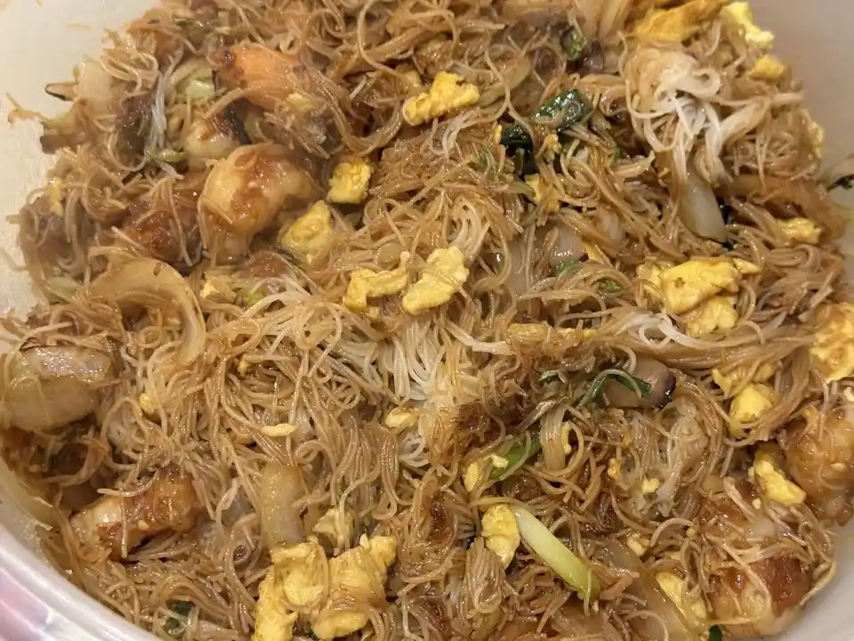 A close-up photo of a bowl of stir-fried rice noodles mixed with shrimp, scrambled egg, green onions, and other vegetables…