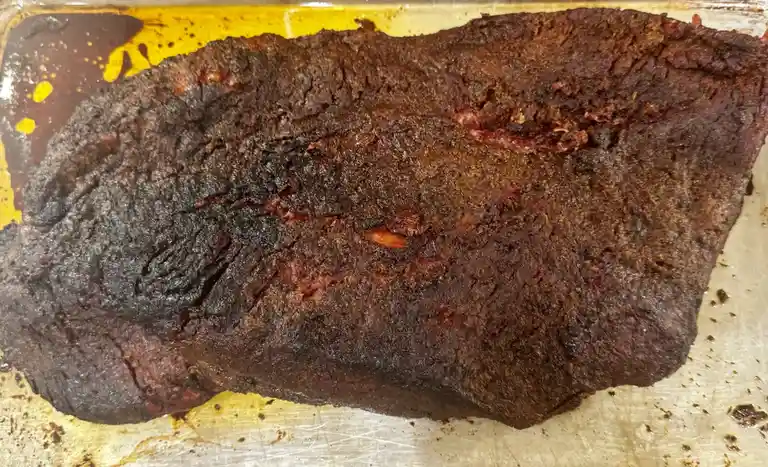 A close-up photo shows a large, heavily charred piece of meat, likely brisket or roast, resting in a yellowish baking pan…