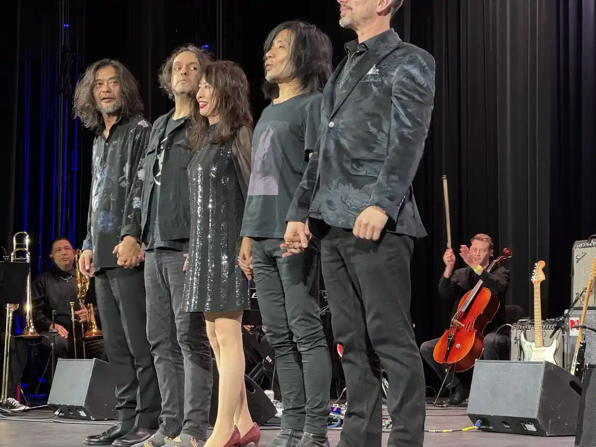 A group of five musicians stand on a stage in front of a black curtain, holding hands and looking upward with expressions of…