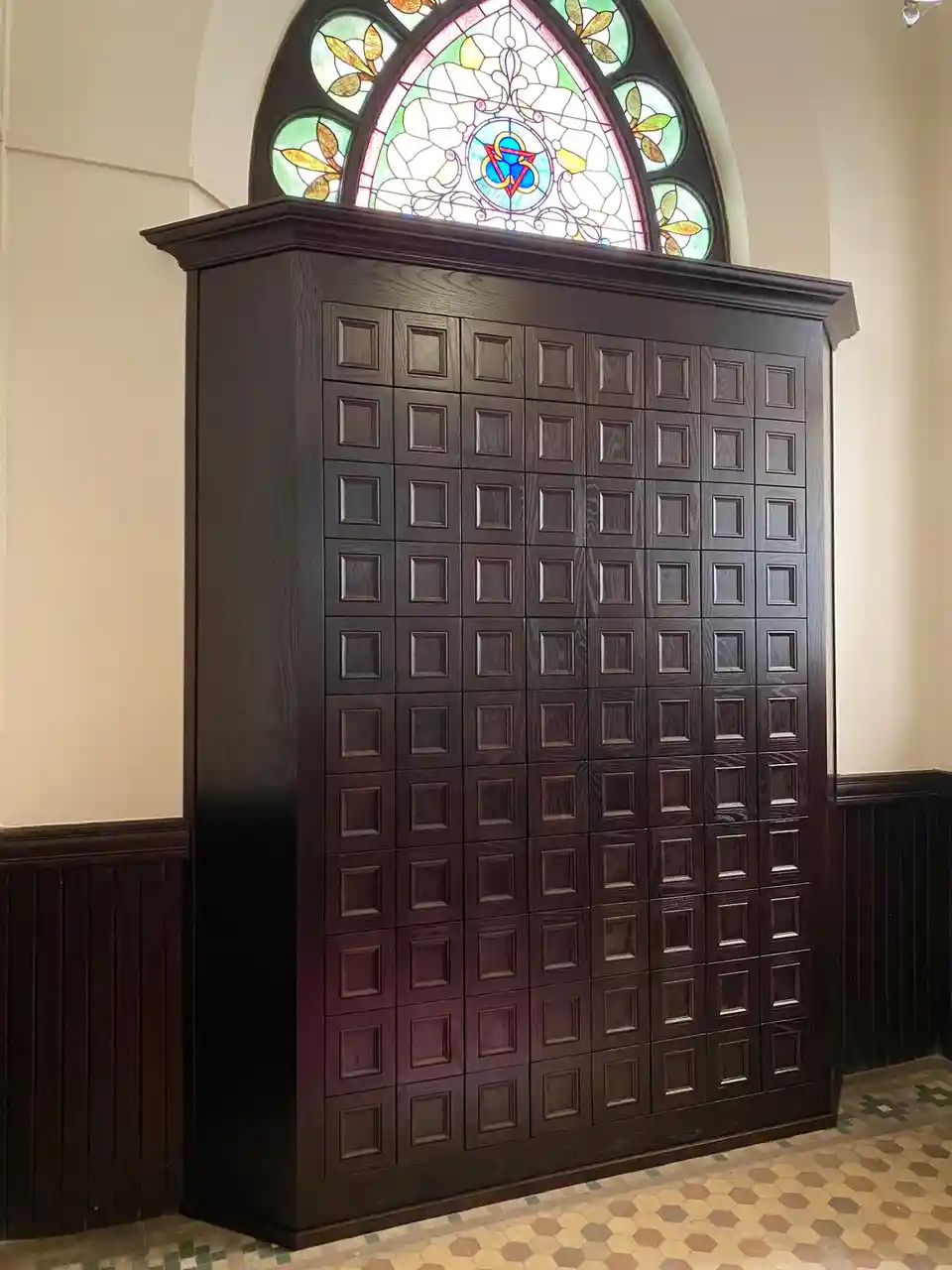 a tall, dark wooden cabinet with a grid of small square compartments stands beneath a stained glass window in a church-like…