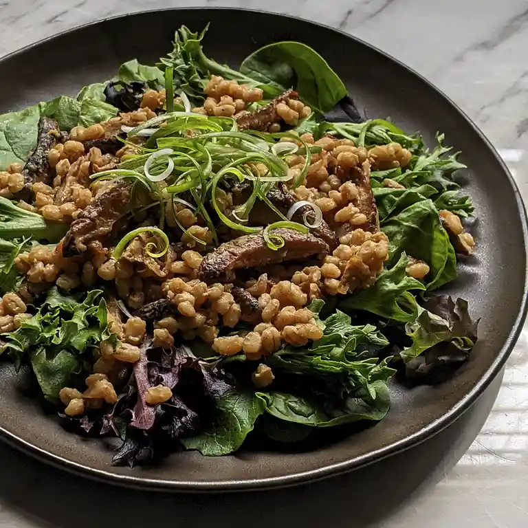 A close-up photo of a plated salad with mixed greens, toasted grains, and strips of cooked meat, garnished with sliced green…