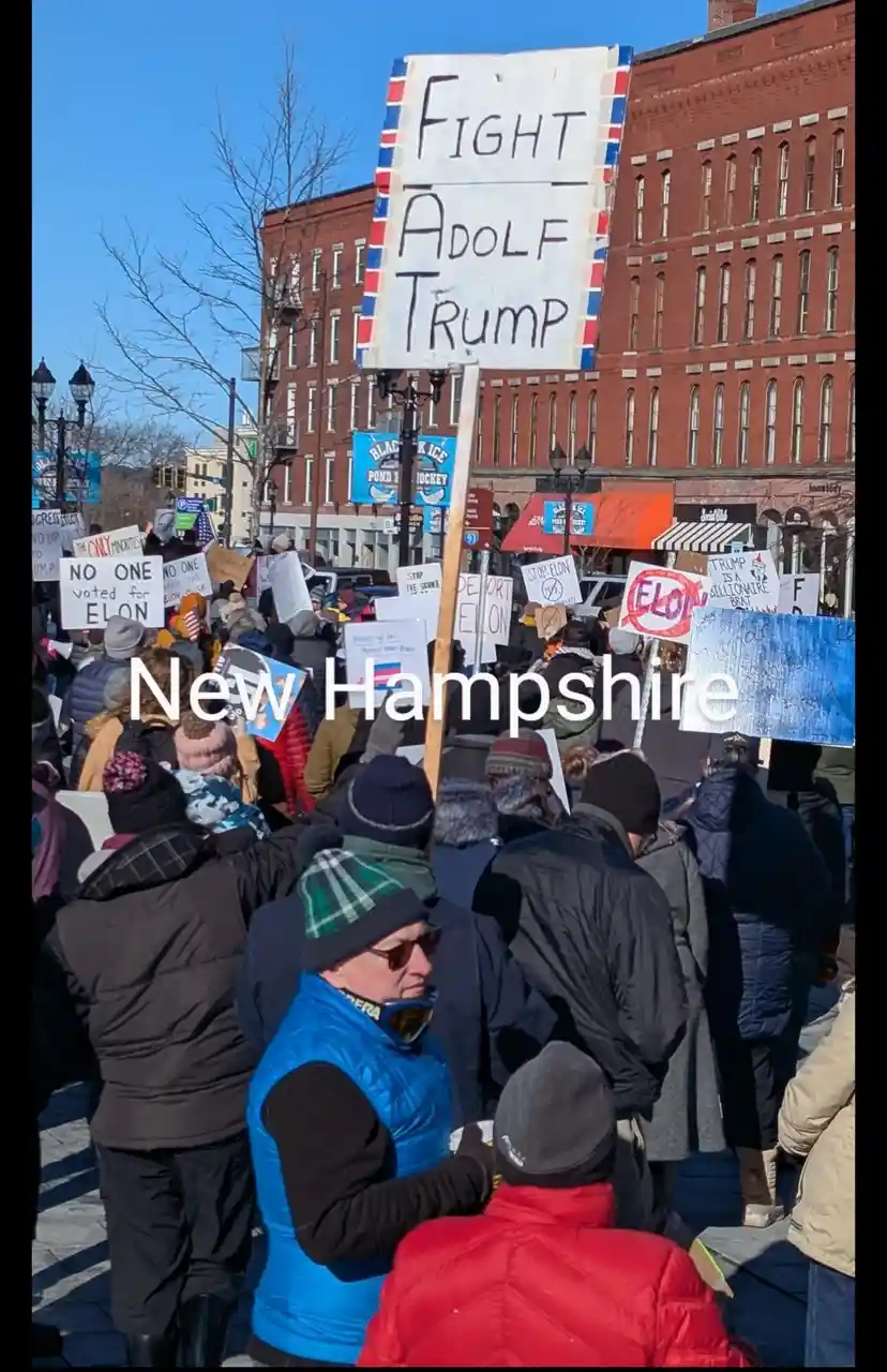 A crowd of protesters in winter coats and hats holds signs at an outdoor rally in New Hampshire. A large handmade sign in…