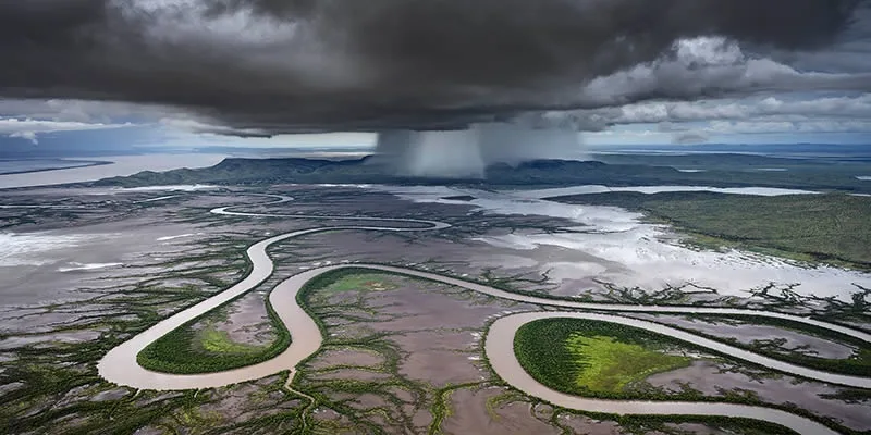 An aerial view shows a wide river meandering through a vast, flat landscape with patches of green vegetation and muddy…