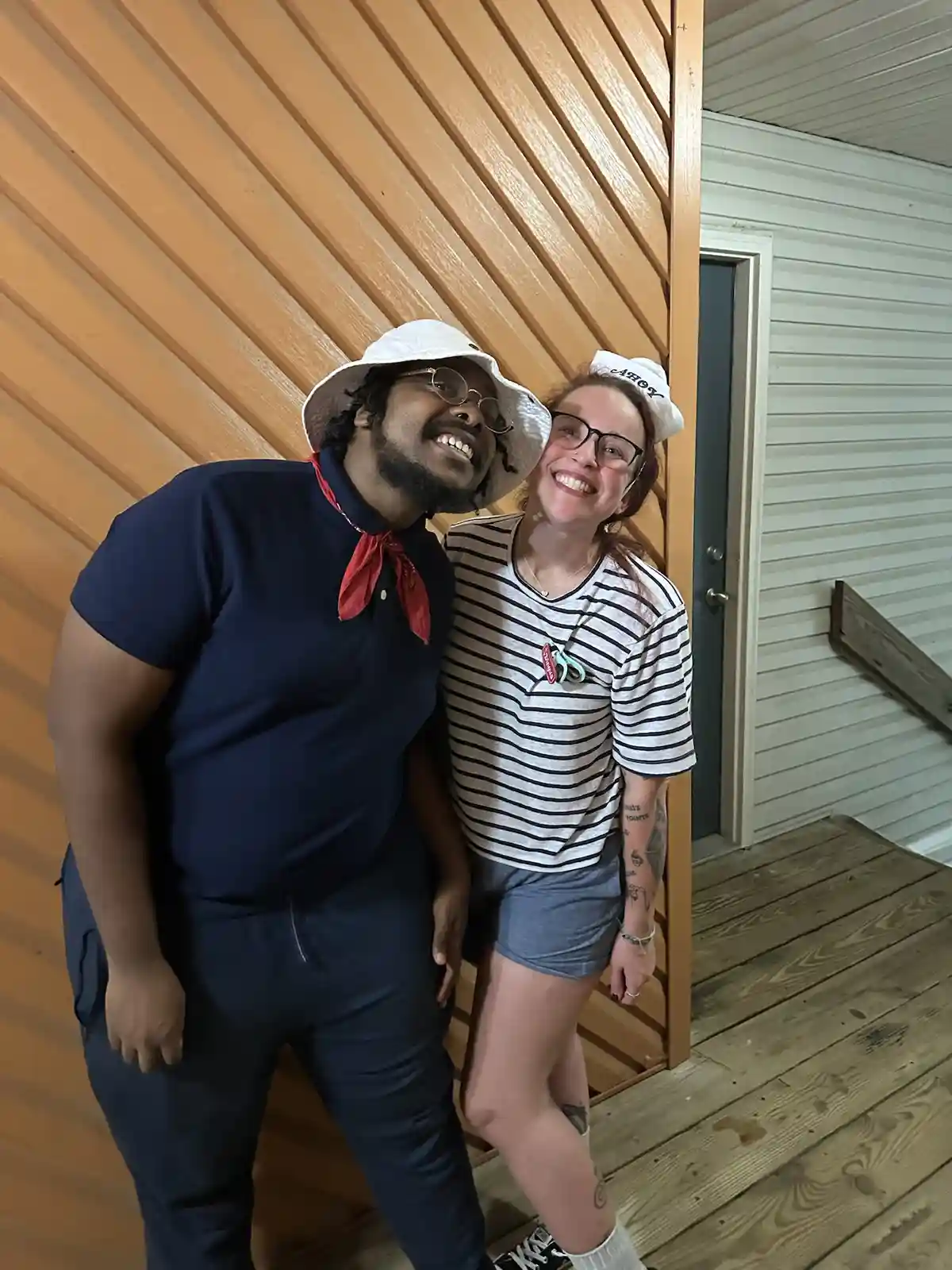 Two smiling people pose together on a wooden porch, both wearing sailor-themed hats and outfits. The person on the left…